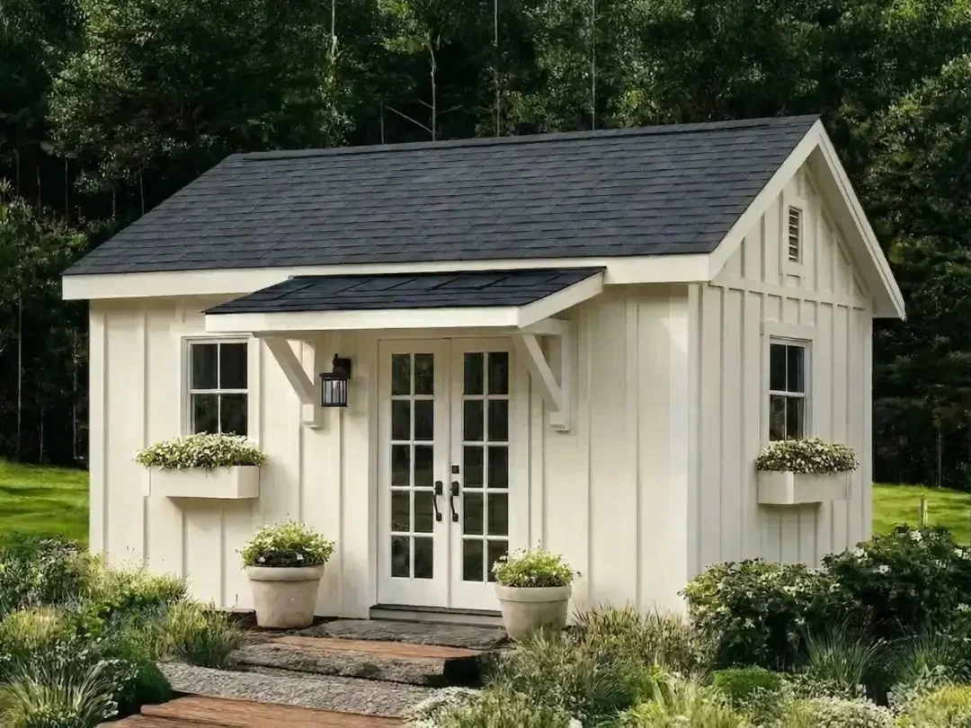 White backyard garden office shed with black roof, French doors, window boxes, and small entry overhang in a landscaped yard.